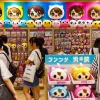 A vibrant retail display of cute chibi-style character goods resembling 'ちいかわ'. The scene is set in a busy Japanese store with plush toys, keychains, and figures neatly organized on shelves. Customers, including Japanese shoppers, are browsing and interacting with the products, creating a lively and engaging atmosphere. The bright colors and playful design of the merchandise capture the fun and charm of these popular items. Horizontal format (16:9).